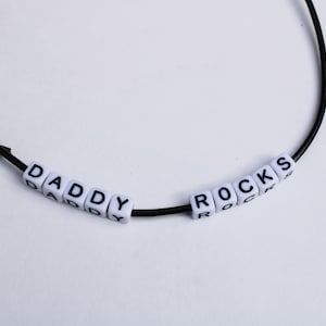 Può includere: Una collana con cordino nero con perline quadrate bianche che compongono la scritta "DADDY ROCKS". Le lettere nere sono centrate sulle perline bianche. La collana è su sfondo bianco.
