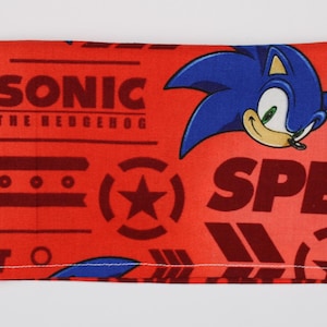 Può includere: Tessuto rosso con un motivo ripetuto con le parole "SONIC THE HEDGEHOG", "SPEED" e "GOTTA GO FAST". Un riccio blu dei cartoni animati è sul lato destro. Il tessuto ha un bordo cucito bianco.