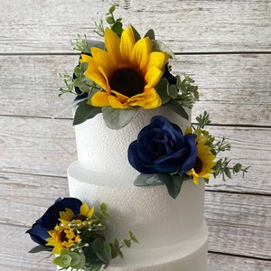 Puede incluir: Un pastel blanco de tres niveles decorado con girasoles amarillos, rosas azul marino y follaje verde. El pastel se encuentra sobre un soporte blanco y ornamentado, perfecto para una boda o evento especial.