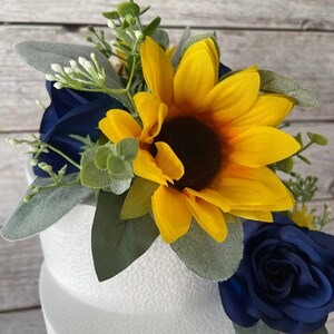 Puede incluir: Arreglo floral decorativo con un gran girasol amarillo con centro marr&oacute;n oscuro, rosas azul marino, hojas verdes y peque&ntilde;os capullos blancos. El arreglo se encuentra sobre un fondo blanco texturizado.