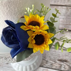 Puede incluir: Arreglo floral con una rosa azul marino y dos girasoles amarillos con centros marrones. Eucalipto verde y peque&ntilde;as flores blancas acent&uacute;an el arreglo, sobre un fondo blanco.