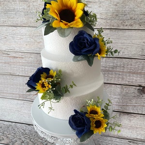 Puede incluir: Un pastel blanco de tres niveles decorado con girasoles amarillos y rosas azul marino. El pastel se encuentra sobre un soporte blanco y ornamentado. La vegetaci&oacute;n acent&uacute;a los arreglos florales, creando un dise&ntilde;o r&uacute;stico y elegante.