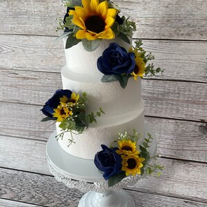 Puede incluir: Un pastel blanco de tres niveles decorado con rosas azul marino, girasoles amarillos y vegetaci&oacute;n. El pastel se encuentra sobre un soporte blanco y ornamentado, perfecto para una boda o evento especial.