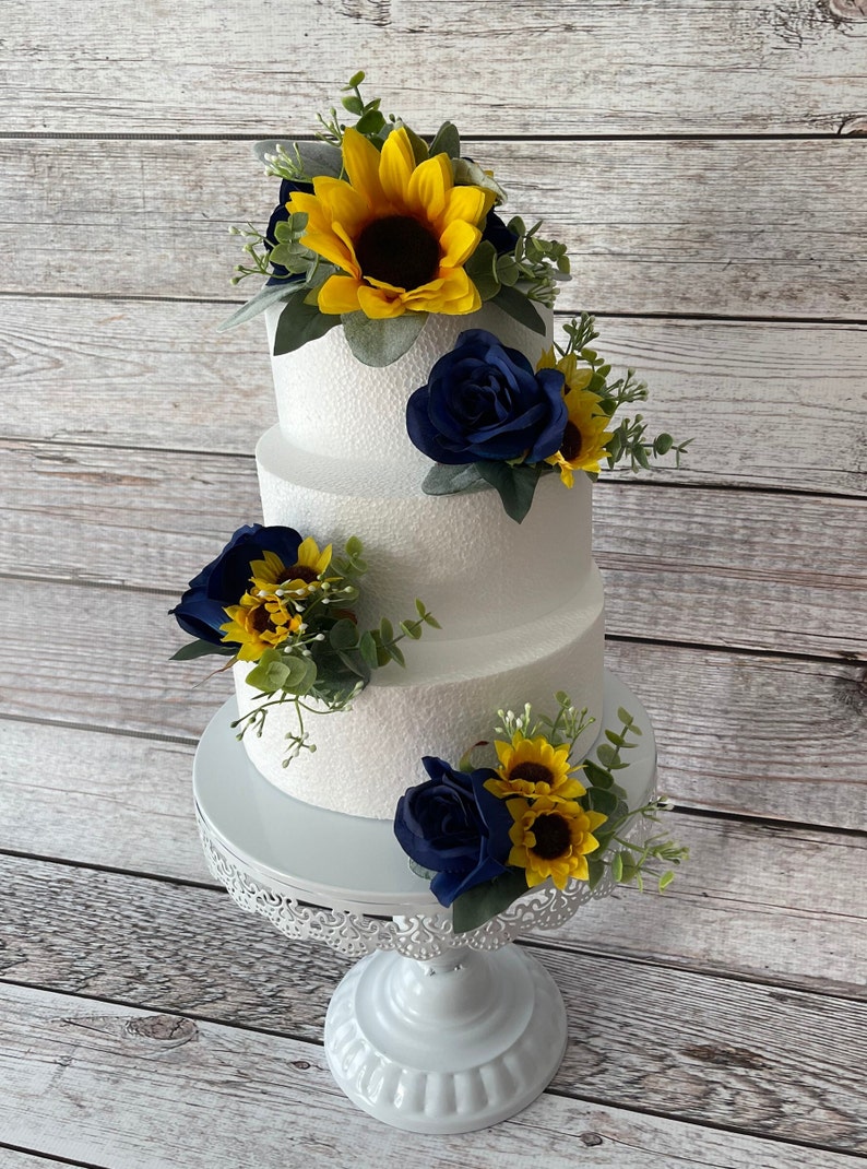 Puede incluir: Un pastel blanco de tres niveles decorado con rosas azul marino, girasoles amarillos y vegetaci&oacute;n. El pastel se encuentra sobre un soporte blanco y ornamentado, perfecto para una boda o evento especial.