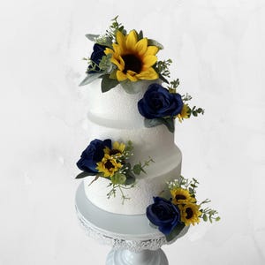 Puede incluir: Un pastel blanco de tres niveles decorado con rosas azul marino, girasoles amarillos y vegetaci&oacute;n. El pastel se encuentra sobre un soporte blanco y ornamentado, creando una pieza central visualmente atractiva.