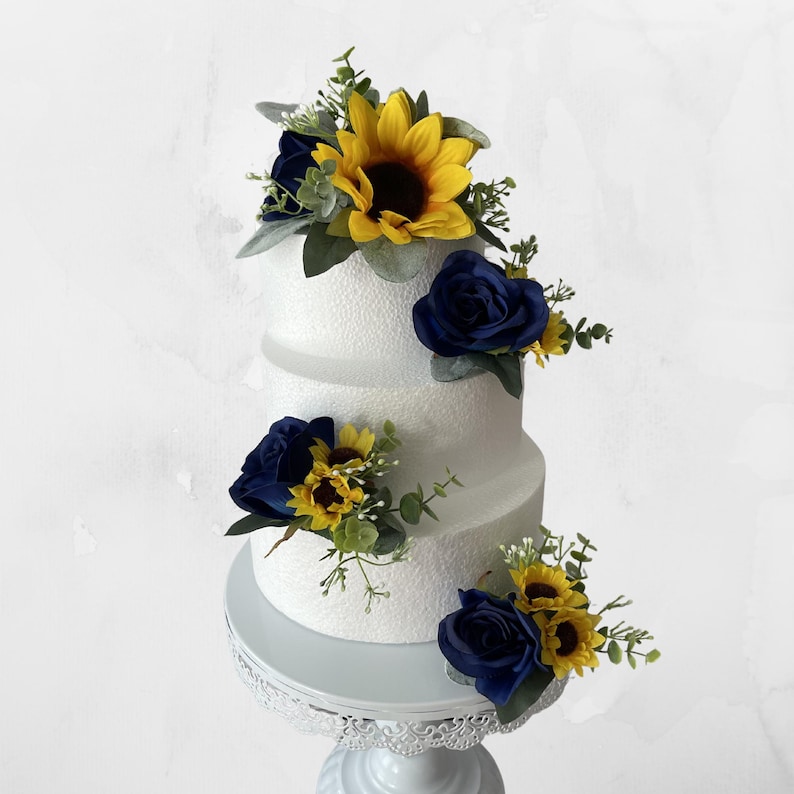 Puede incluir: Un pastel blanco de tres niveles decorado con rosas azul marino, girasoles amarillos y vegetaci&oacute;n. El pastel se encuentra sobre un soporte blanco y ornamentado, creando una pieza central visualmente atractiva.