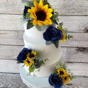 Puede incluir: Un pastel blanco de tres niveles decorado con rosas azul marino, girasoles amarillos y vegetaci&oacute;n. El pastel se encuentra sobre un soporte para pasteles blanco y ornamentado. Las flores est&aacute;n dispuestas en racimos alrededor del pastel.