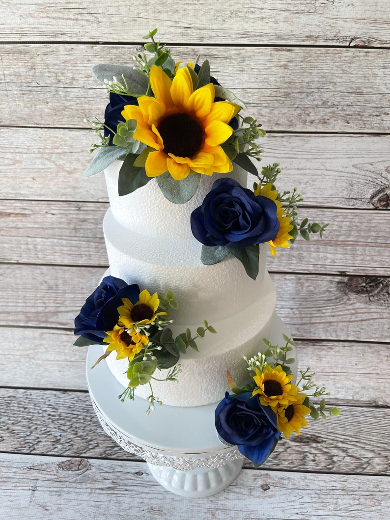 Puede incluir: Un pastel blanco de tres niveles decorado con rosas azul marino, girasoles amarillos y vegetaci&oacute;n. El pastel se encuentra sobre un soporte para pasteles blanco y ornamentado. Las flores est&aacute;n dispuestas en racimos alrededor del pastel.