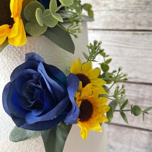 Puede incluir: Primer plano de un arreglo floral con una rosa azul oscuro, girasoles amarillos y follaje verde. El arreglo est&aacute; sobre un fondo blanco, con una superficie de madera visible. Es probable que las flores sean artificiales.