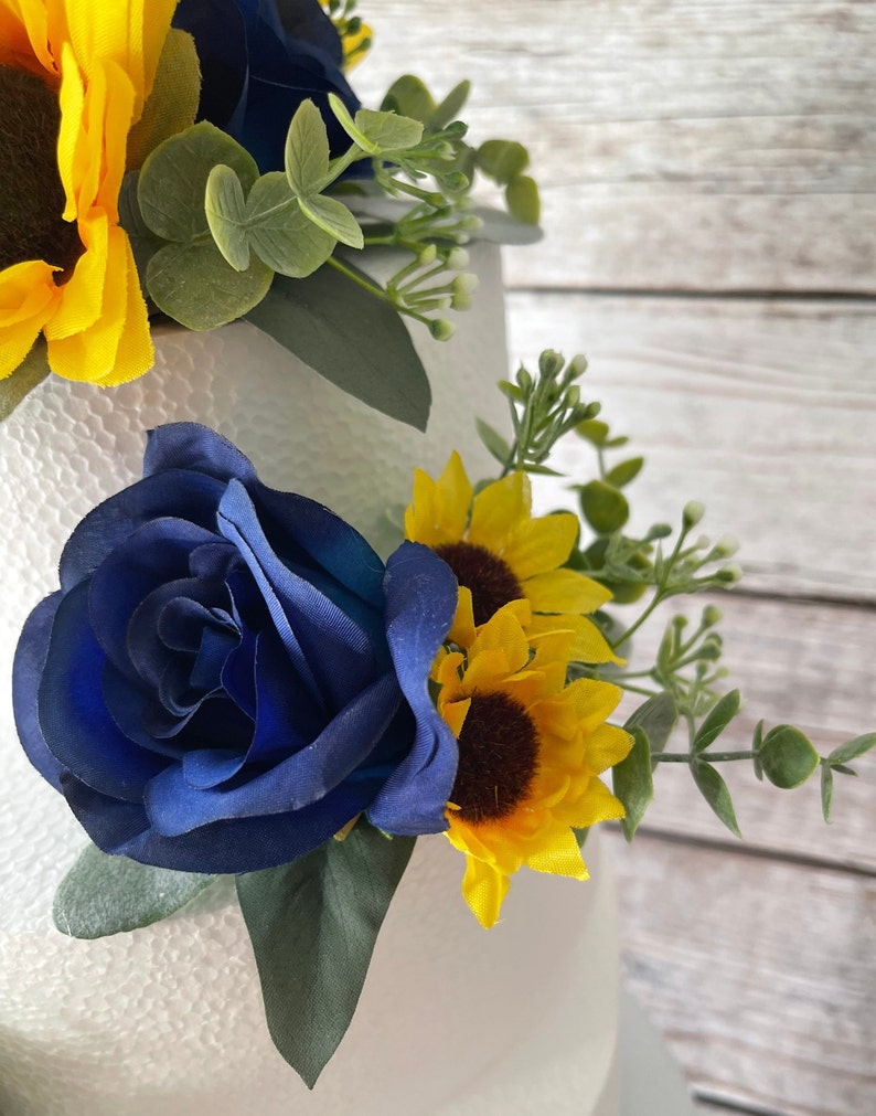 Puede incluir: Primer plano de un arreglo floral con una rosa azul oscuro, girasoles amarillos y follaje verde. El arreglo est&aacute; sobre un fondo blanco, con una superficie de madera visible. Es probable que las flores sean artificiales.