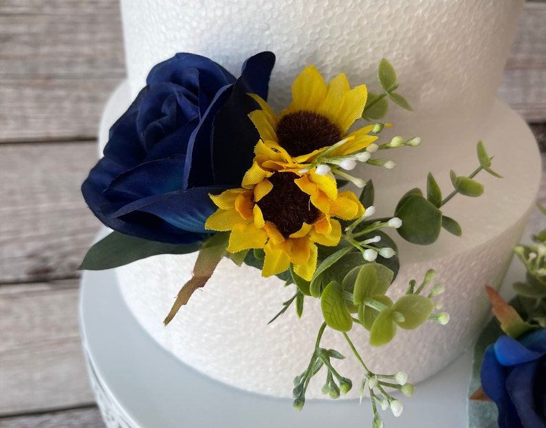 Puede incluir: Una rosa azul marino y dos girasoles amarillos con centros marrones se acent&uacute;an con hojas verdes y peque&ntilde;os capullos blancos. El arreglo floral es probablemente para una boda o un evento especial.