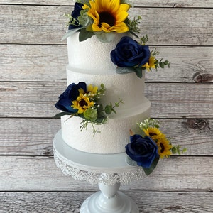 Puede incluir: Un pastel blanco de tres niveles decorado con girasoles, rosas azul marino y vegetaci&oacute;n. El pastel se encuentra sobre un soporte blanco y ornamentado para pasteles, sobre un fondo de madera clara. El pastel es una pieza central para una boda o celebraci&oacute;n.