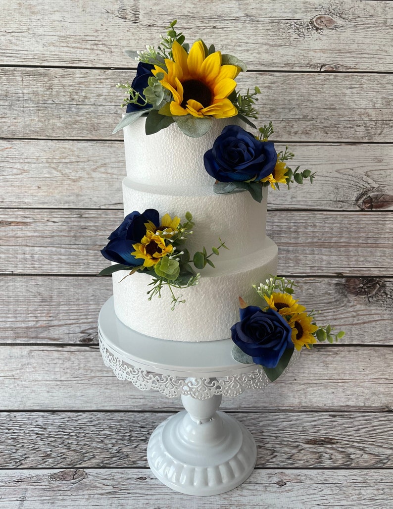 Puede incluir: Un pastel blanco de tres niveles decorado con girasoles, rosas azul marino y vegetaci&oacute;n. El pastel se encuentra sobre un soporte blanco y ornamentado para pasteles, sobre un fondo de madera clara. El pastel es una pieza central para una boda o celebraci&oacute;n.