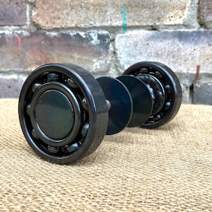 May include: A detailed view of a black bearing, partially taken apart, revealing ball bearings and dark grey metal parts. The bearing rests on a textured, beige surface, with a brick wall in the background.