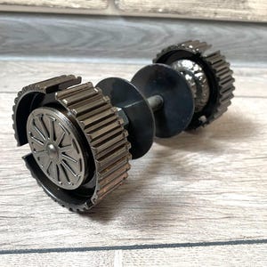 Pode incluir: Um haltere feito à mão, feito de engrenagens e componentes de metal reciclados. O haltere apresenta uma haste central com placas circulares e extremidades em forma de engrenagem, mostrando uma estética industrial única. O metal tem um esquema de cores prateado e preto.