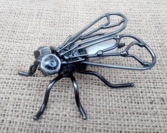 May include: A handcrafted metal sculpture of a fly. The insect is constructed from various metal pieces, including nuts, bolts, and wire, with a white cylindrical element in the body. The wings are made of curved wire, and the legs are curved metal rods.