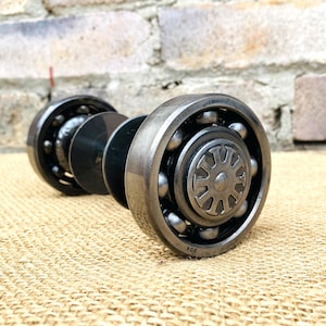 May include: A metallic industrial-style door knob constructed from two circular ball bearings connected by a central rod. The outer rings of the bearings are polished steel, while the center features a decorative, gear-like circular metal insert.