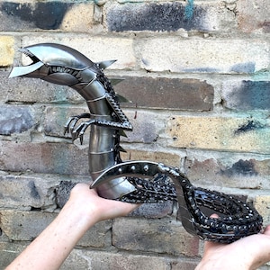 Könnte beinhalten: Eine Metallskulptur eines Wesens mit länglichem Kopf, scharfen Zähnen und einem segmentierten Körper. Die Figur besitzt einen geschwungenen Schwanz aus dunklen, texturierten Kettengliedern und glatte, polierte Metallplatten am Rumpf und Kopf.