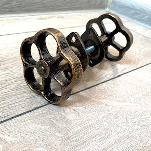 Può includere: Un paio di pomelli per mobili in bronzo in stile antico. Ogni pomello presenta una maniglia circolare a forma di fiore con finitura anticata. I pomelli sono montati su una base rettangolare con fori per le viti.