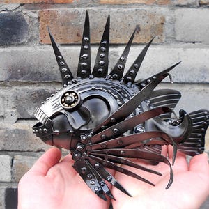 Op de afbeelding: Een handgemaakte metalen sculptuur van een vis met een uniek industrieel ontwerp. De sculptuur heeft scherpe, puntige vinnen en stekels, en een gedetailleerde kop in mechanische stijl. De vis wordt in de handen van een persoon gehouden, wat de grootte en ingewikkelde constructie laat zien.