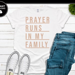 Puede incluir: Camiseta blanca con el texto "PRAYER RUNS IN MY FAMILY" en color marrón claro. La camiseta se combina con vaqueros azules y zapatillas blancas. La imagen está sobre una superficie de madera blanca.