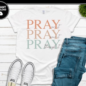 Op de afbeelding: Wit T-shirt met de woorden "PRAY on it, PRAY over it, PRAY through it" in perzik, lichtbruin en blauwgroen. Het shirt wordt gecombineerd met een blauwe jeans en witte sneakers. Een zwarte cirkel met "INSTANT DOWNLOAD" staat linksboven.