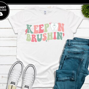 Könnte beinhalten: Weißes T-Shirt mit dem Text "KEEP ON BRUSHIN'" in Retro-Schrift. Das Design beinhaltet einen lächelnden Zahn, einen Blitz und Sterne in Rosa-, Grün- und Koralltönen. Das Shirt wird mit blauen Jeans und weißen Turnschuhen kombiniert.