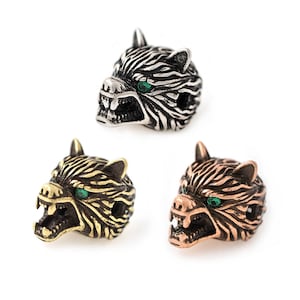 May include: Three metal wolf head beads in silver, gold, and copper tones. Each bead features detailed fur texture, open mouths with visible teeth, and green gemstone eyes. The beads are designed for jewelry making or crafting.