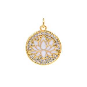 Puede incluir: Colgante dorado con un diseño de flor de loto blanca en el centro. La flor está rodeada por un círculo de pequeños cristales transparentes. El colgante tiene un pequeño lazo en la parte superior para una cadena.