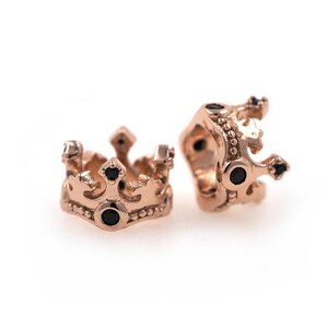 May include: Two gold-toned crown-shaped beads with black gemstones. The beads are designed to be used in jewelry making.