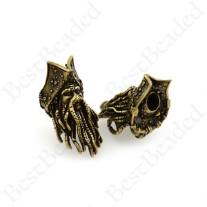 Puede incluir: Dos cuentas de metal de color dorado antiguo con un diseño detallado de calavera de pirata. Una cuenta muestra la calavera con barba y sombrero, mientras que la otra cuenta muestra la parte posterior de la calavera.