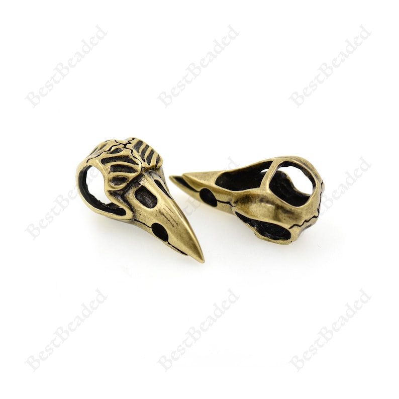 May include: Two antique gold-toned metal bird skull charms. The charms are detailed with intricate bone-like designs.
