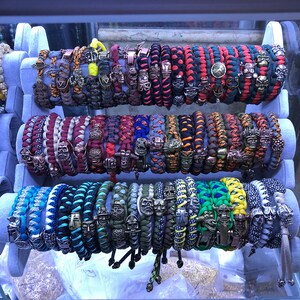 May include: A collection of colorful paracord bracelets with skull and other metal charms. The bracelets are displayed on a white stand.
