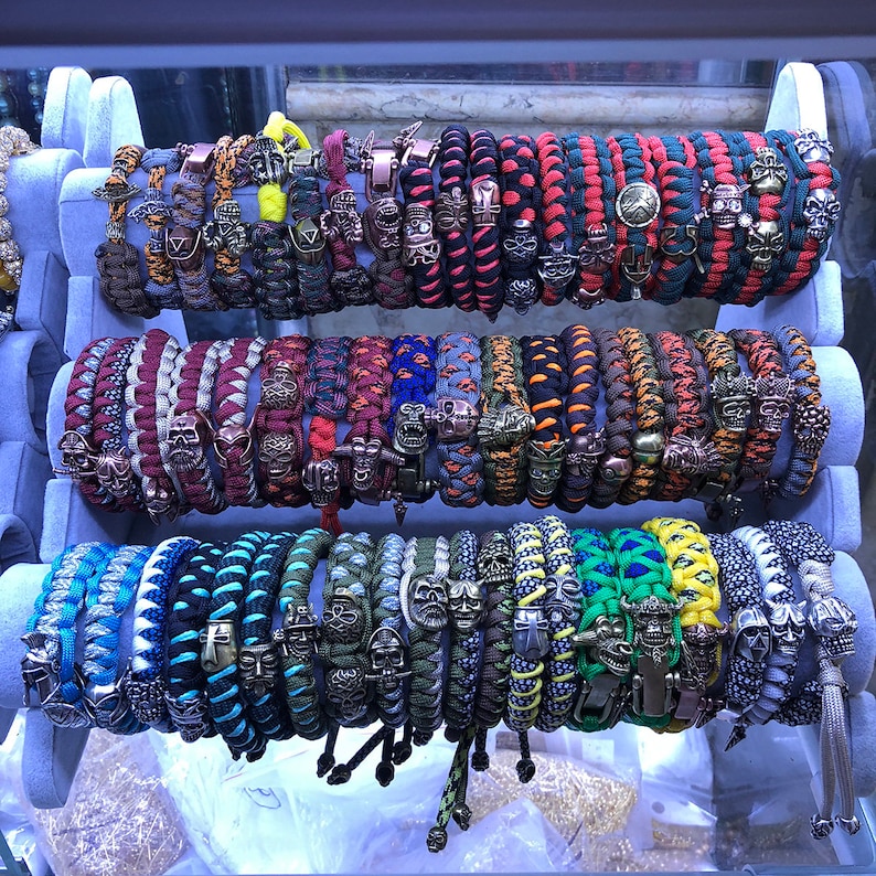 May include: A collection of colorful paracord bracelets with skull and other metal charms. The bracelets are displayed on a white stand.