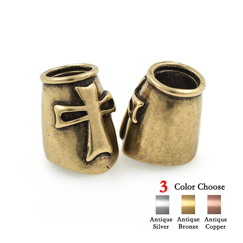 May include: Two antique bronze-colored metal beads with a cross design. The beads have a cylindrical shape with a cross on the front. The image also shows color options: antique silver, antique bronze, and antique copper.