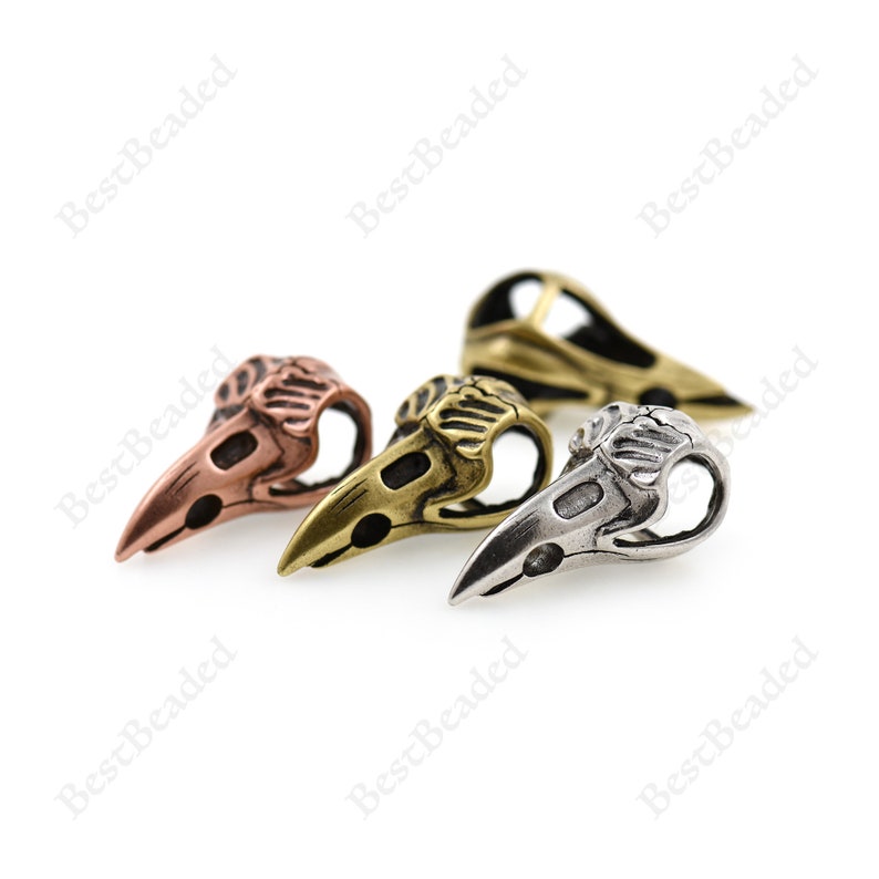 May include: Three metal bird skull beads in different colors: copper, brass, and silver. The beads are detailed and have a realistic bird skull design.