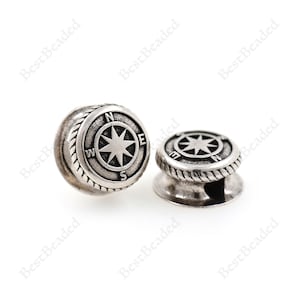 May include: Two silver-toned metal beads with a compass design. Each bead features a raised compass rose with the letters N, S, E, and W around the edge. The beads have a rope-like design around the edge.