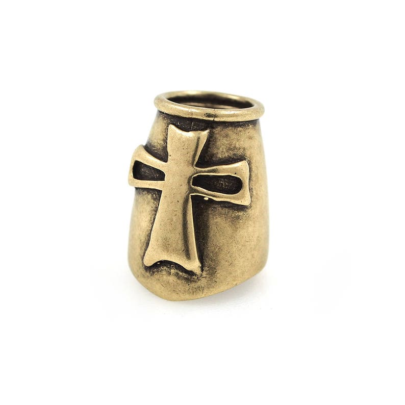 May include: A bronze-colored metal bead with a cross design. The bead has a cylindrical shape with a flared top and a cross cutout on the front. The bead is likely used for jewelry making or other crafts.