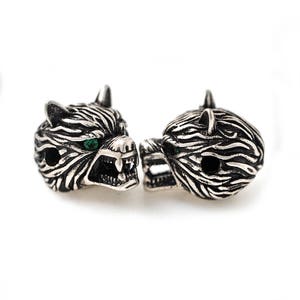 May include: Two silver-toned wolf head beads with green eyes. The beads have detailed fur texture and open mouths, revealing teeth. The beads are likely for jewelry making or crafting.