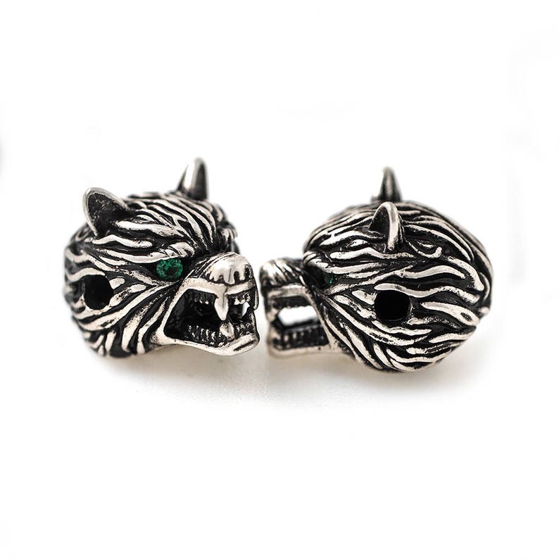 May include: Two silver-toned wolf head beads with green eyes. The beads have detailed fur texture and open mouths, revealing teeth. The beads are likely for jewelry making or crafting.