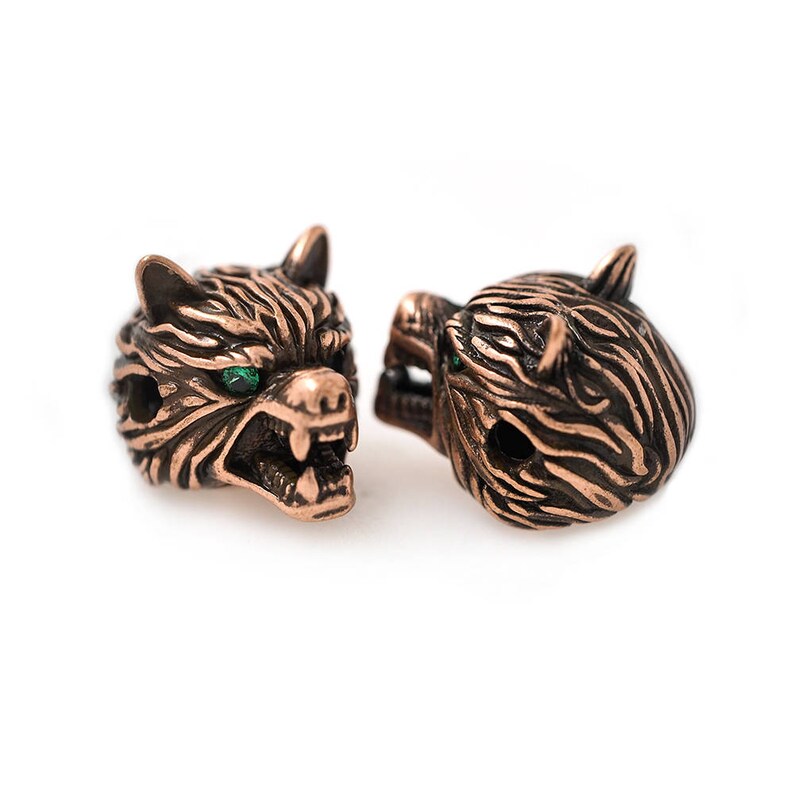 May include: Two bronze-colored wolf head beads with green gem eyes. The beads are detailed with carved fur and open mouths, showcasing sharp teeth. The beads are designed for jewelry making or crafting.