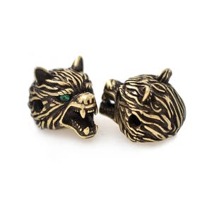 May include: Two bronze-colored tiger head beads with green eyes. The beads have detailed stripes and open mouths, showcasing sharp teeth. These decorative beads are ideal for jewelry making or crafting projects.