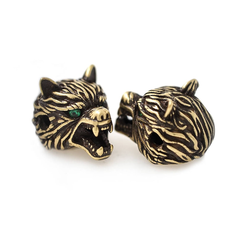 May include: Two bronze-colored tiger head beads with green eyes. The beads have detailed stripes and open mouths, showcasing sharp teeth. These decorative beads are ideal for jewelry making or crafting projects.