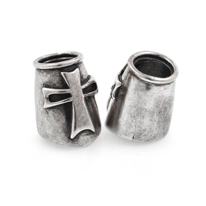 May include: Two silver-toned metal beads with a cross design. The beads have a cylindrical shape with a slightly flared top. The cross is prominently displayed on the front of each bead, adding a religious or symbolic element.