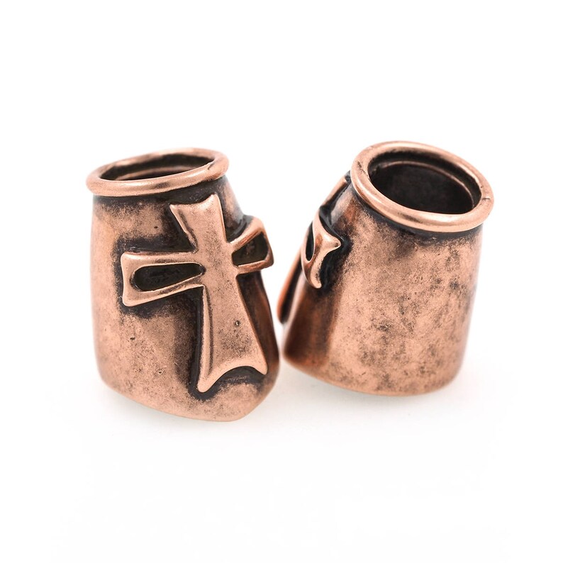 May include: Two copper-colored metal beads with a cross design. The beads have a cylindrical shape with a slightly flared top and a detailed cross on the front. The finish has a rustic, aged appearance.
