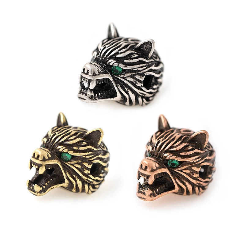 May include: Three metal wolf head beads in silver, gold, and copper tones. Each bead features detailed fur texture, open mouths with visible teeth, and green gemstone eyes. The beads are designed for jewelry making or crafting.