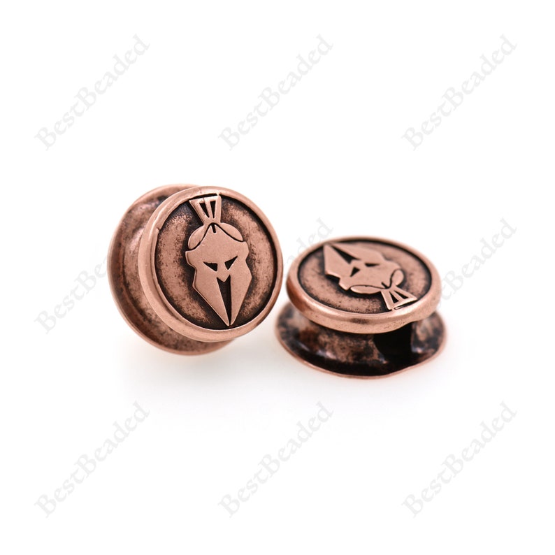 May include: Two copper-colored metal beads with a raised design of a Spartan helmet. The beads are round and have a smooth surface.