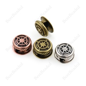 May include: Three compass-shaped metal toggles in different finishes: antique copper, antique brass, and silver. Each toggle features a compass rose with the letters N, S, E, and W around the star.