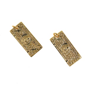 May include: Two gold-colored pendant earrings shaped like a $100 bill. The earrings are detailed with the image of Benjamin Franklin and the text "ONE HUNDRED DOLLARS".