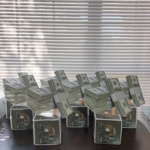 May include: A row of eight tissue boxes decorated with stacks of money. Each box is made of cardboard and has a clear plastic window. The boxes are arranged in a row on a dark surface.